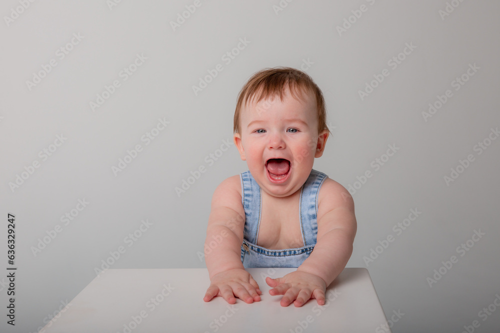 A charming little baby boy in a blue jumpsuit sits on a white background, looks at the camera and smiles. A charming little baby boy in a blue jumpsuit sits on a white background, looks at the camera and smiles.