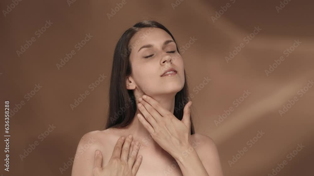The young woman enjoys her smooth, clean silky skin, touches the neck, shoulders and chest. Portrait of a seminude smiling woman in the studio on a brown background. The young woman enjoys her smooth, clean silky skin, touches the neck, shoulders and chest. Portrait of a seminude smiling woman in the studio on a brown background.
