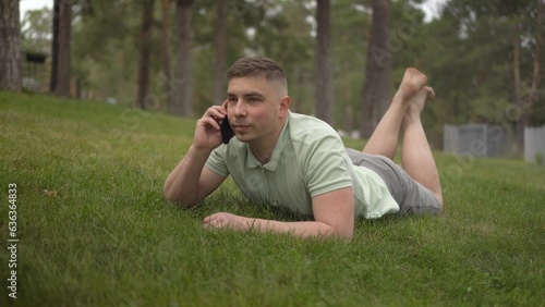 A young man lies on the grass and talks on the phone. A student in the park rests on the grass with communicates by cellular connection.