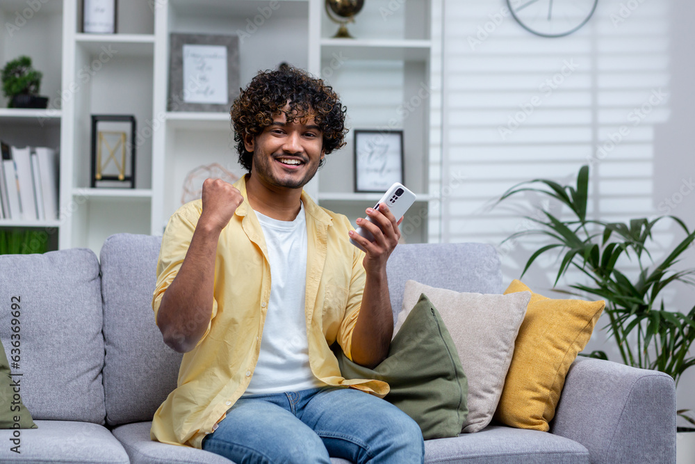 Portrait of young successful man at home on sofa, winner smiling and looking at camera holding phone in hands using game app, holding hand up successful gesture of triumph. Portrait of young successful man at home on sofa, winner smiling and looking at camera holding phone in hands using game app, holding hand up successful gesture of triumph.