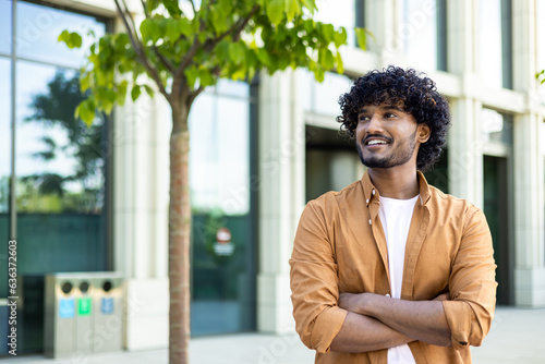 Portrait of a successful young male designer, engineer standing outside an office center, crossing his arms on his chest and looking to the side with a smile