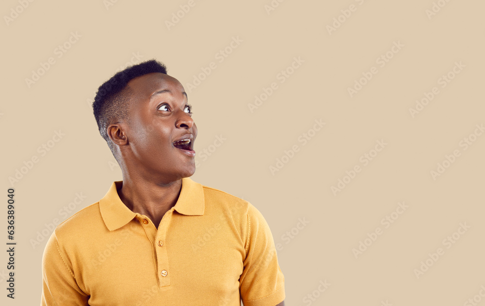 Fototapeta premium Young positive WOW African American man looks up with enthusiastic emotions when he sees unexpected promotional offer or surprise with huge discounts for shopping, stands in beige studio. Copy space