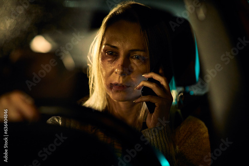A young Caucasian woman, bearing the distressing signs of domestic abuse with prominent bruises and blackened eyes, speaks on her phone as she navigates her car through the nighttime streets.