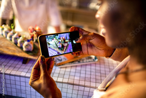 Young african woman taking a photo with her smart phone of the sushi a chef prepared in a sushi bar photo