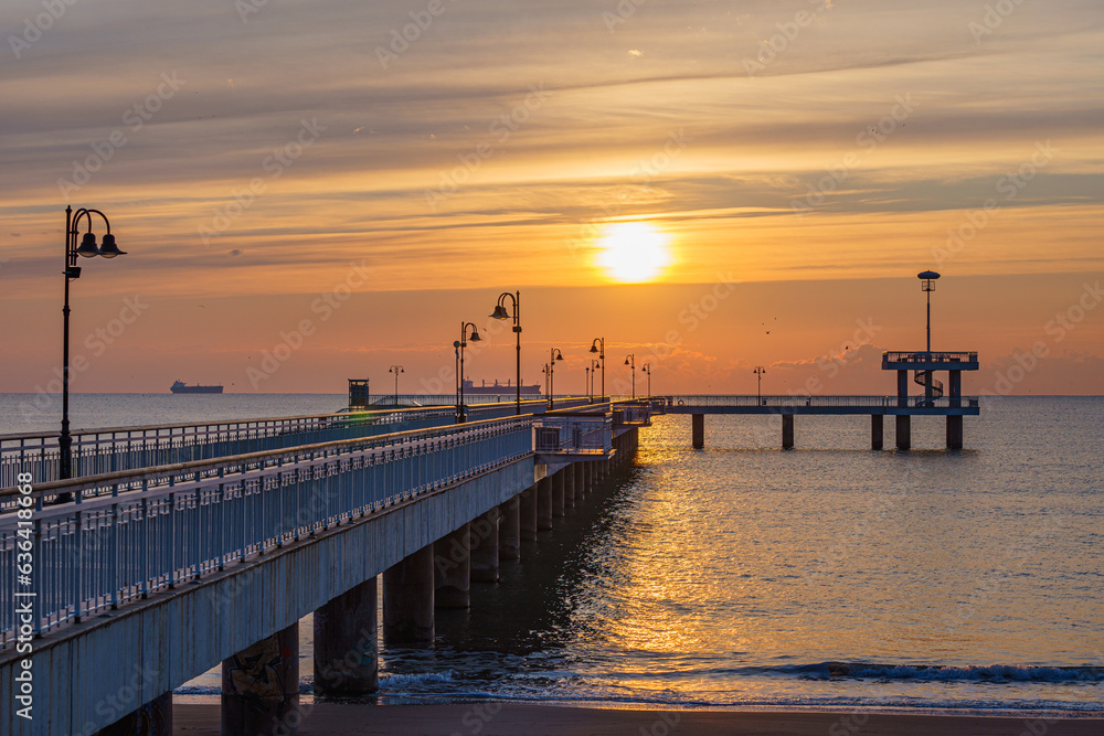 Obraz premium Bridge at the beach extending into the ocean with distant boats under a bright orange sun at sunset