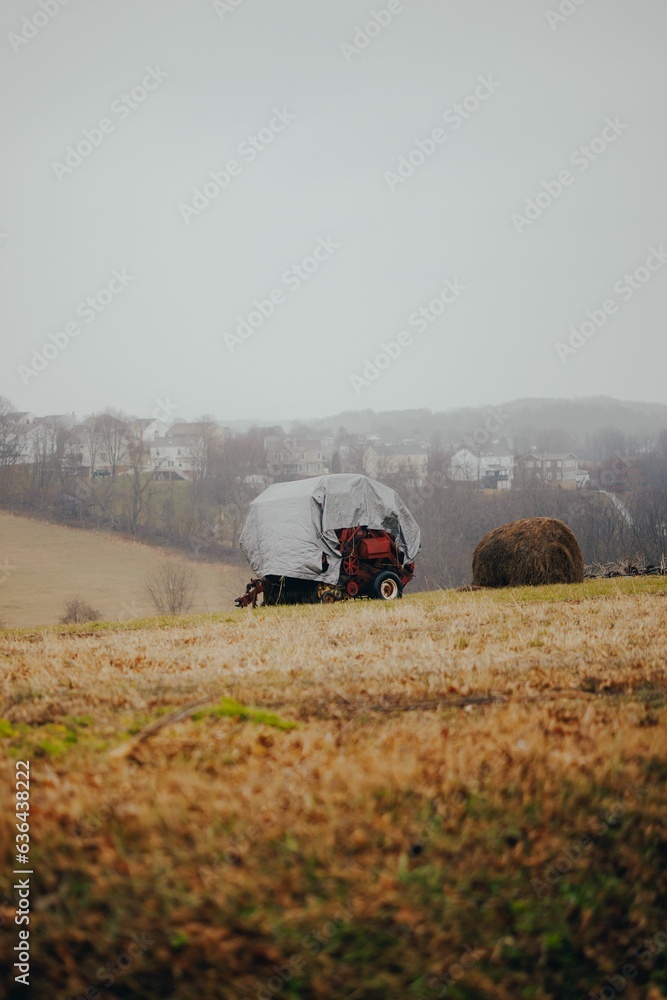 Fototapeta premium Tractor covered with a weatherproof cover near a hay bale on a misty day