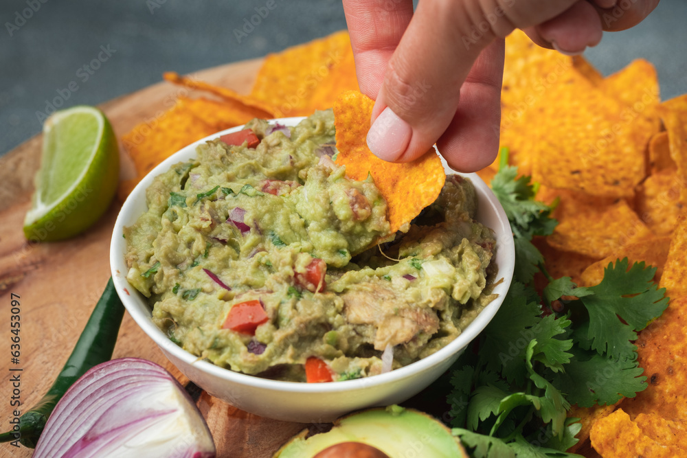 Woman hand dip nachos into tasty guacamole sauce on the wooden background. Tortilla chips next to guacamole ingredients: avocado, cilantro, onion, hot pepper and lime. Traditional Mexican food