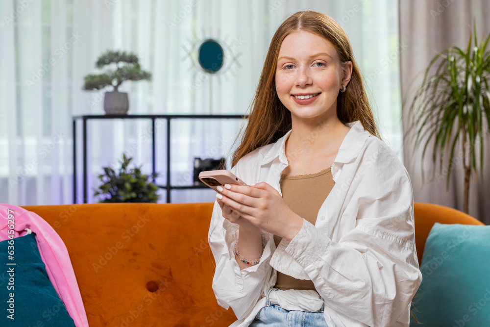 Teenager smiling girl sitting on sofa uses mobile phone at home living room looking at camera. Child texting share messages content on smartphone social media applications online watching relax movie Teenager smiling girl sitting on sofa uses mobile phone at home living room looking at camera. Child texting share messages content on smartphone social media applications online watching relax movie