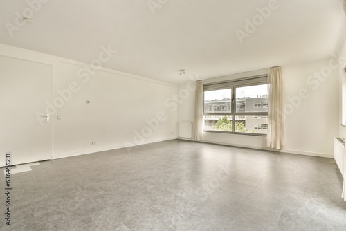 an empty living room with white walls and grey flooring  there is a large window overlooking out onto the street