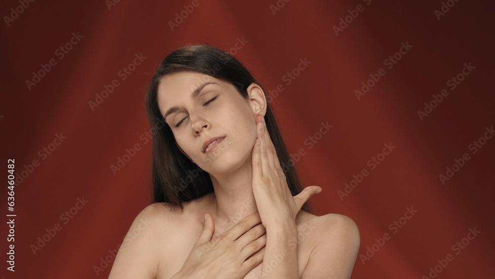 Fototapeta premium The young woman enjoys her smooth, clean silky skin, touches the neck and chest. Portrait of a seminude smiling woman in the studio on a red background. The concept of beauty, cosmetology, skin care.