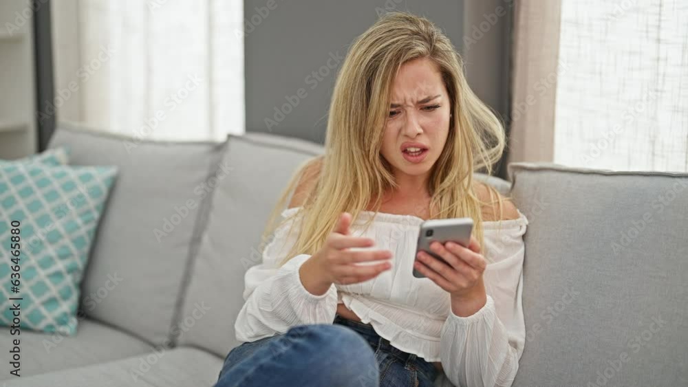 Young blonde woman using smartphone sitting on sofa looking upset at home Young blonde woman using smartphone sitting on sofa looking upset at home
