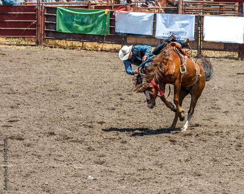 A rodeo cowboy is riding a bucking bronco. He is in an arena with dirt flying from the kicking horse. There is fence railing in the background. The cowboy is wearing a black vest and blue shirt.