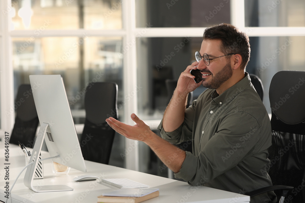 © New Africa - Happy man using modern computer while talking on smartphone at white desk in office
