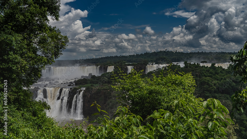 Waterfall landscape. Streams of water cascade from the ledges. Lush tropical vegetation all around. Clouds in the blue sky. Iguazu Falls. Brazil.