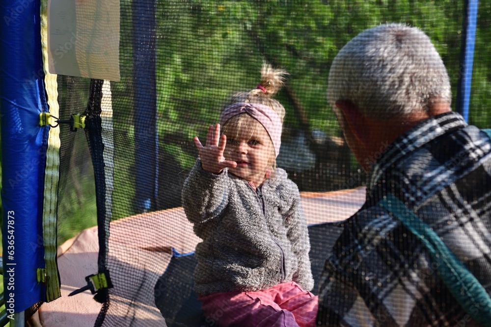 A little girl runs around in a trampoline and kicks a ball with her grandfather