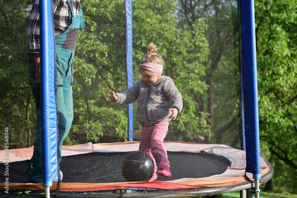 A 2-year-old girl jumps and kicks a ball on a trampoline in the summer