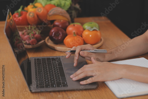 A young woman with a beautiful face in a blue shirt with long hair eating fruit sitting inside the kitchen at home with a laptop and notebook for relaxation, Concept Vacation.