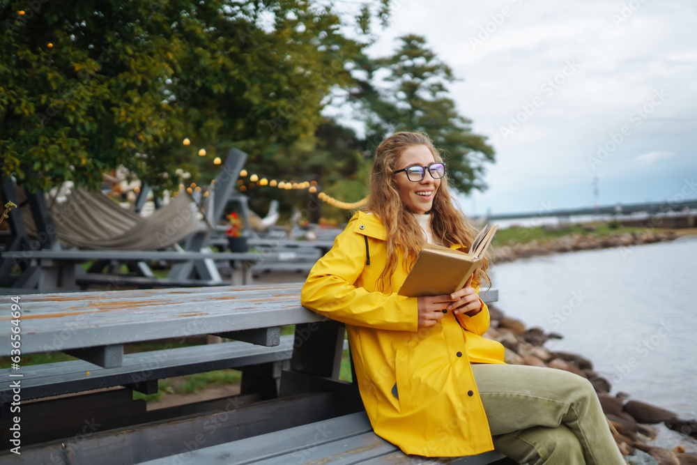 Fototapeta premium Smiling young woman sits on a bench and reads a book, enjoys the view of the lake. The concept of relaxation, enjoyment, solitude with nature.