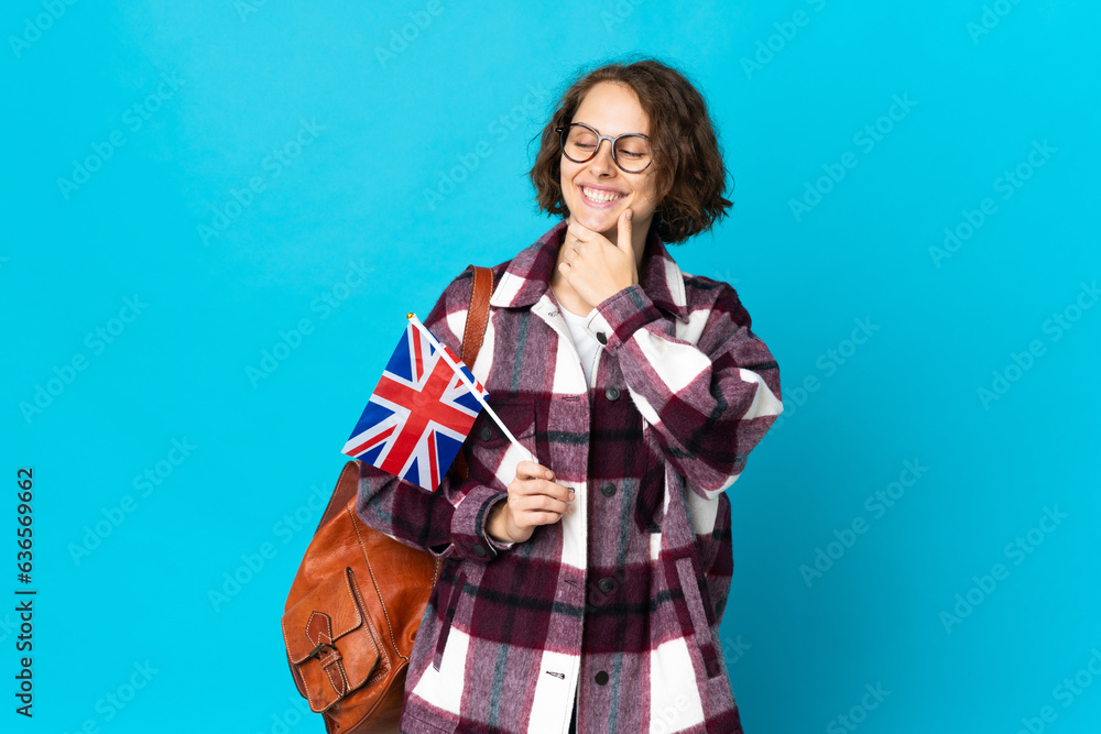 Young English woman holding an United Kingdom flag isolated on blue background looking to the side and smiling