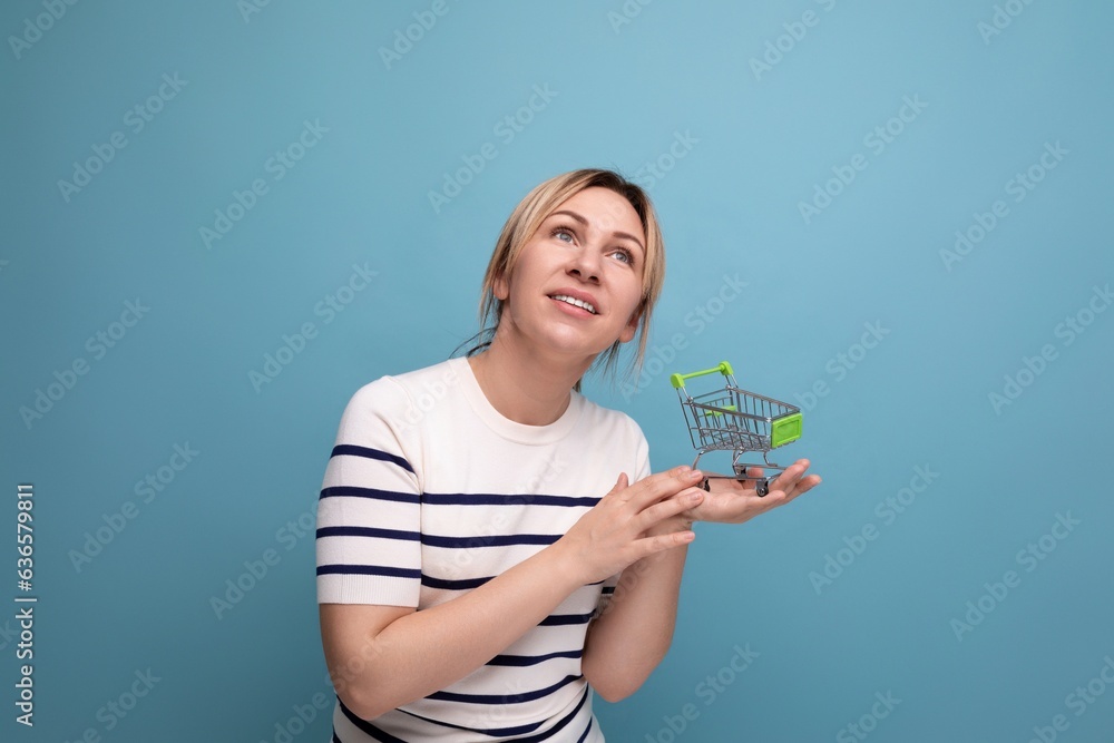 horizontal photo of young cute casual shopaholic woman holding supermarket shopping trolley on blue background with copy space horizontal photo of young cute casual shopaholic woman holding supermarket shopping trolley on blue background with copy space