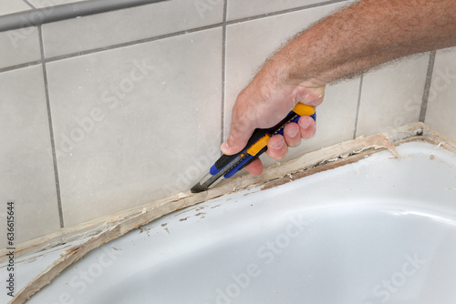 Closeup of plumber hand removing old dirty silicone from bathtube in a washroom, cutting of silicone glue using cutter, bathroom renovation