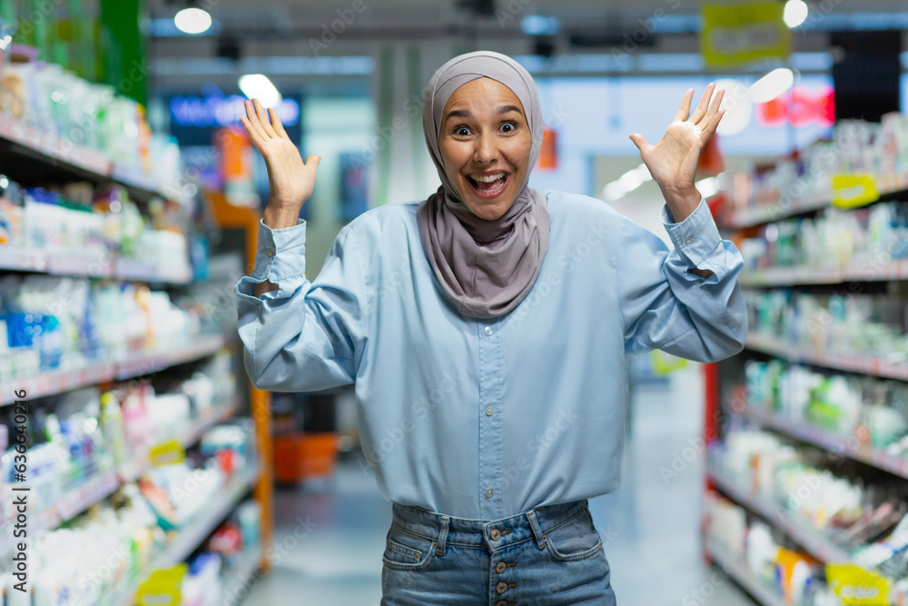 Portrait of a young Muslim woman in a hijab standing in a supermarket in the department of household chemicals. Shows with his hands, chooses a product, looks into the camera, poses, advertises.