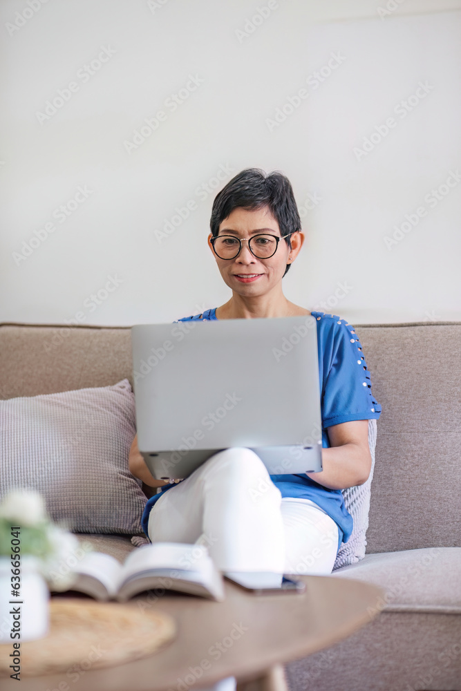 Happy Asian 60s retired woman enjoys social media on laptop while relaxing in living room. Happy Asian 60s retired woman enjoys social media on laptop while relaxing in living room.