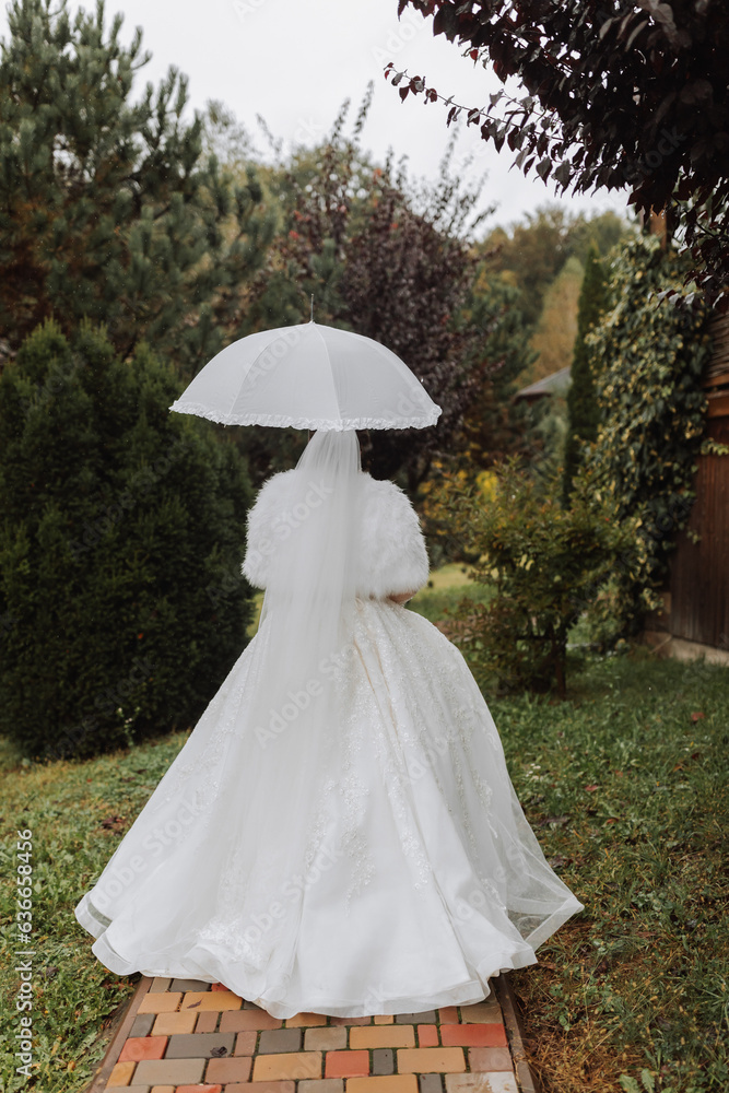 Beautiful stylish bride with a white umbrella, walks in the rain in the park, wedding photo session in the rain. A model with an umbrella in a lush wedding dress