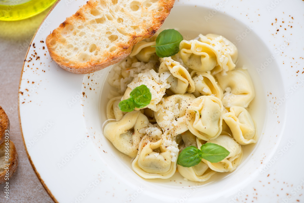 Tortellini in bouillon served with grated parmesan, green basil and ciabatta in a beige plate, close-up, selective focus