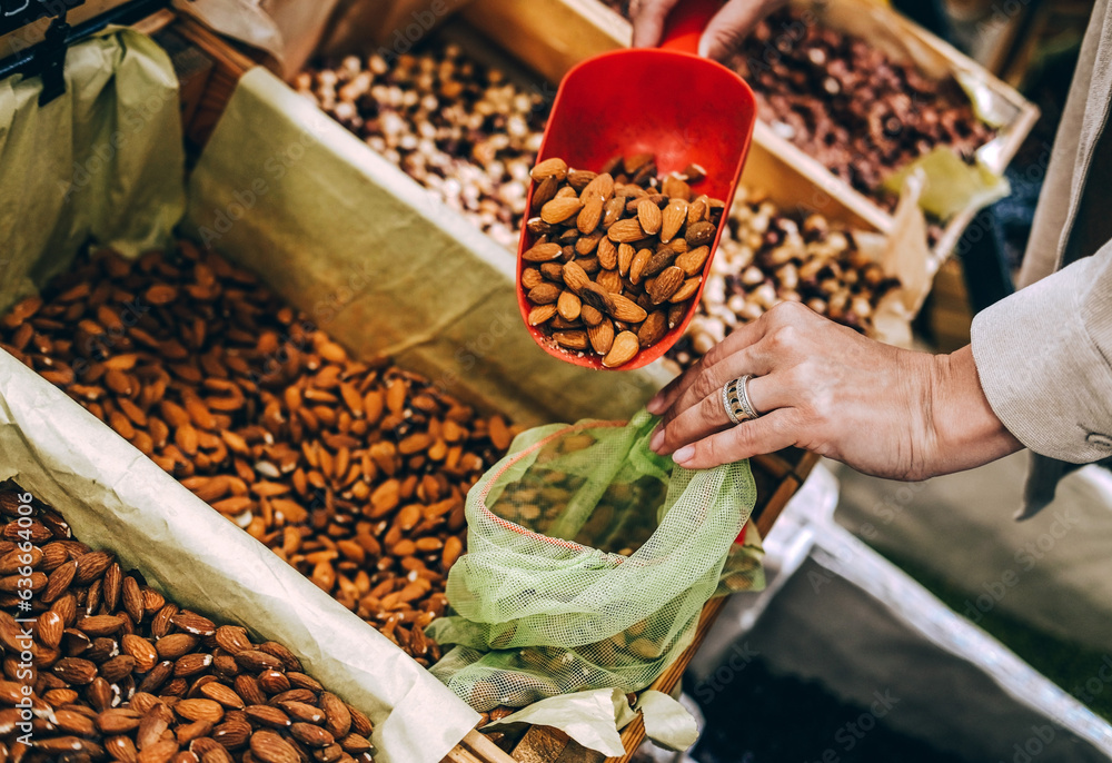 © dvulikaia - Close-up of  young woman pouring almonds into  mesh reusable bag at market.