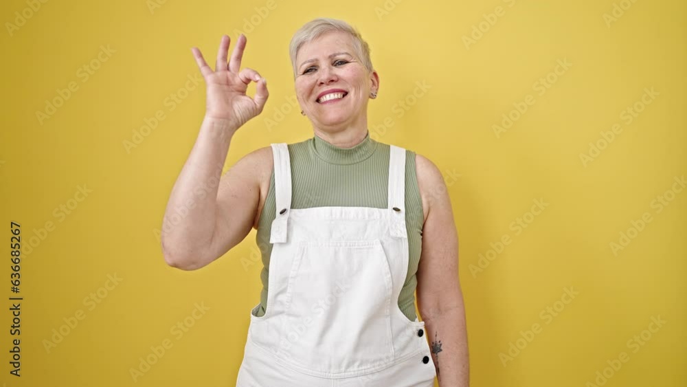 Middle age grey-haired woman smiling with ok gesture over isolated yellow background Middle age grey-haired woman smiling with ok gesture over isolated yellow background