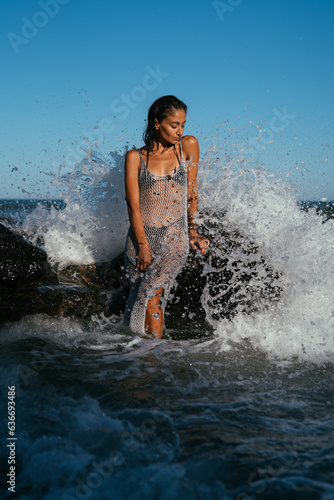 A beautiful wet brunette girl in a silvery net stands in the water on the seashore