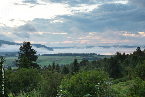 High angle view of a hill covered in trees, the Baie-Saint-Paul valley and the St. Lawrence River seen from route 138 during a beautiful foggy summer sunrise, Charlevoix, Quebec, Canada