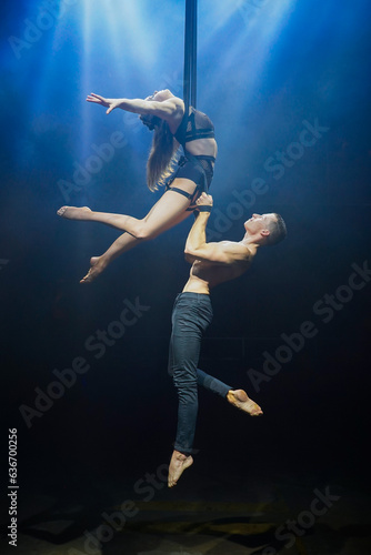 Fototapeta Naklejka Na Ścianę i Meble -  An aerial straps duo performs dangerous tricks mid-air, illuminated by strong blue and white lights against a dark background, clad in minimal athletic attire, showcasing a blend of power and grace