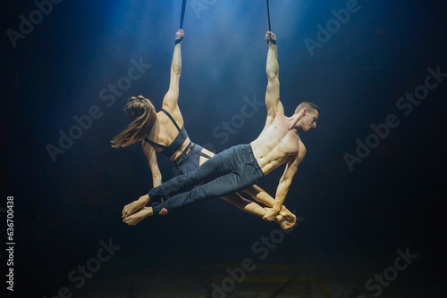 Aerial straps duo performance: a man and woman execute graceful acrobatic feats in mid-air against a black background, dressed in black and illuminated by a white-blue glow
