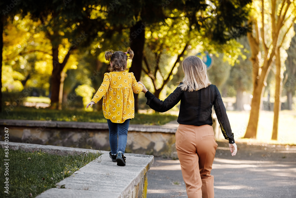 Obraz premium Mothers day. Happy young mom is holding by hand and walking with adorable little daughter at autumn park. Mom with kid are having fun, playing at weekend. Outdoors vacation activity. Children's day.
