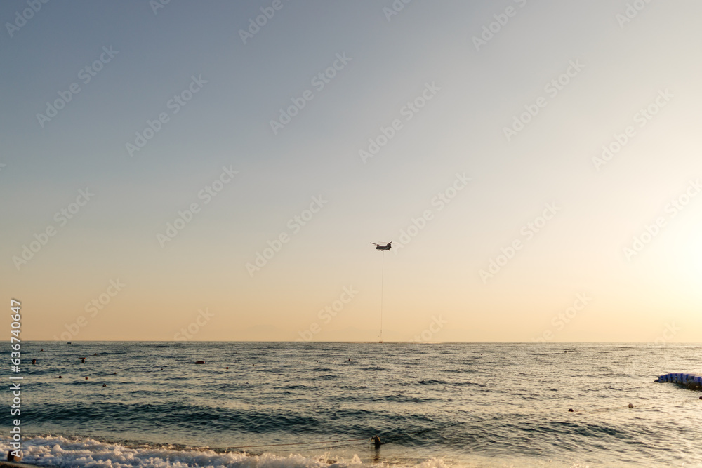 Sunrise on the ocean. Beautiful sea view from the shore. The helicopter will deliver water from the sea to put out the fire in the wild forest. Forest fire in Kemer, Antalya, Türkiye. Sunrise on the ocean. Beautiful sea view from the shore. The helicopter will deliver water from the sea to put out the fire in the wild forest. Forest fire in Kemer, Antalya, Türkiye.