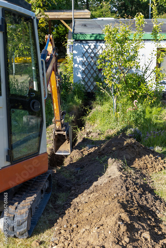A mini excavator is digging a trench in front of a garage for an electrical ground cable