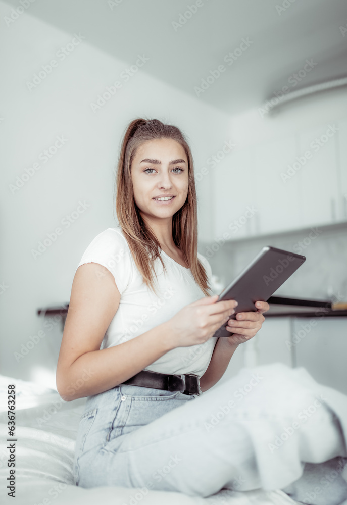 Young smiling woman using tablet while sitting on bed in bright room Young smiling woman using tablet while sitting on bed in bright room