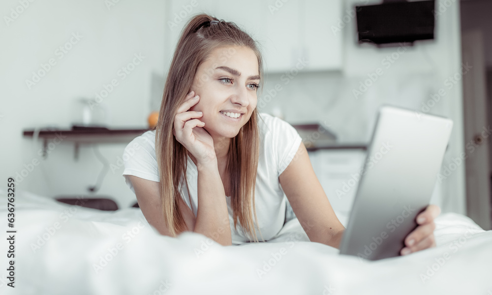 Young smiling woman looking at tablet screen while lying on bed in bright room Young smiling woman looking at tablet screen while lying on bed in bright room