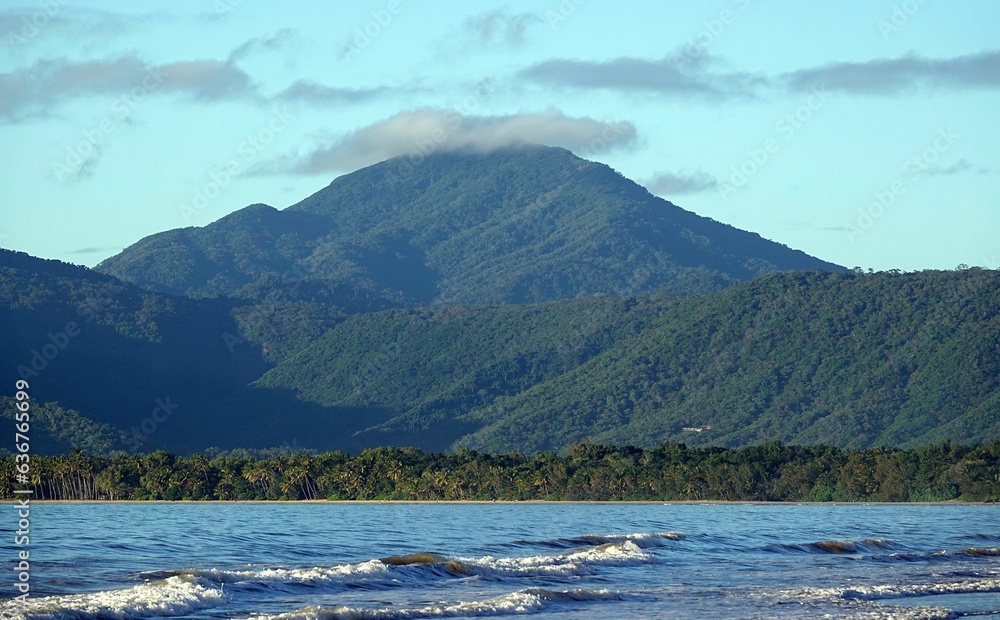 a mountain near a beach with waves crashing towards it and trees and hills
