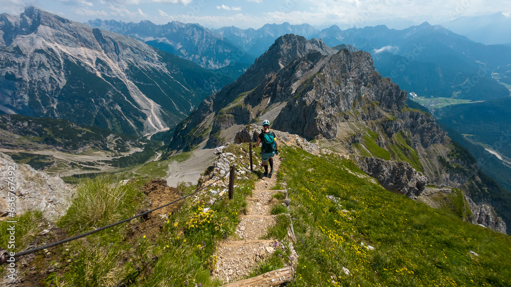A female hiker descends a narrow mountain path along the Mittenwalder Höhenweg, surrounded by dramatic alpine peaks and lush green meadows under a bright summer sky. A female hiker descends a narrow mountain path along the Mittenwalder Höhenweg, surrounded by dramatic alpine peaks and lush green meadows under a bright summer sky.
