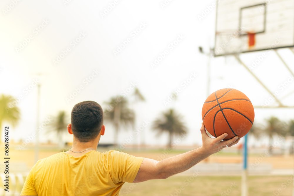 Fototapeta premium Young man with his back to the camera holding the basketball with one hand on an outstretched arm. Blurred basketball hoop background. Triumph concept.