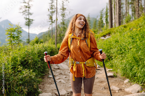 Stylish tourist woman with a yellow backpack and hiking equipment walks along a hiking trail against the backdrop of mountain scenery. The concept of travel, vacation. Active lifestyle.