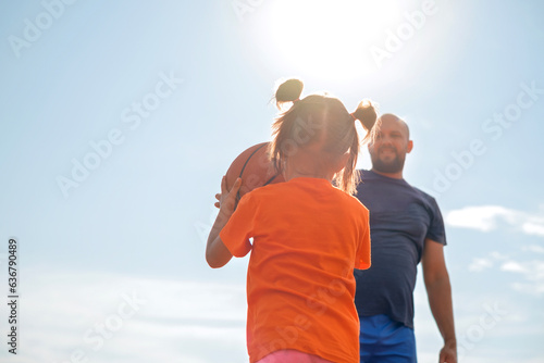 Funny little kid playing basketball with his father, rear view against the background of the sky and the sun. Dad teaches daughter to play with a ball