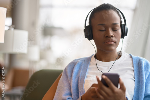 Calm African Black hipster teen girl student refugee sitting indoors, wearing headphones using mobile phone, listening music mix online for relaxation with eyes closed, meditating. Copy space. photo