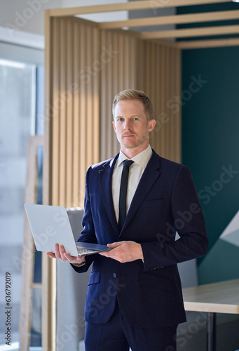 Confident young adult professional business man company leader, male ceo executive manager wearing formal suit and tie looking at camera holding laptop standing in office. Vertical portrait.