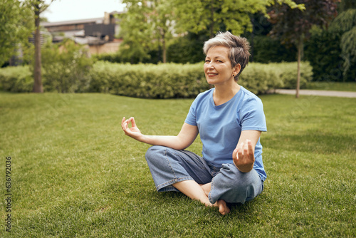 Senior woman in lotus pose sitting on green grass while doing yoga at city park