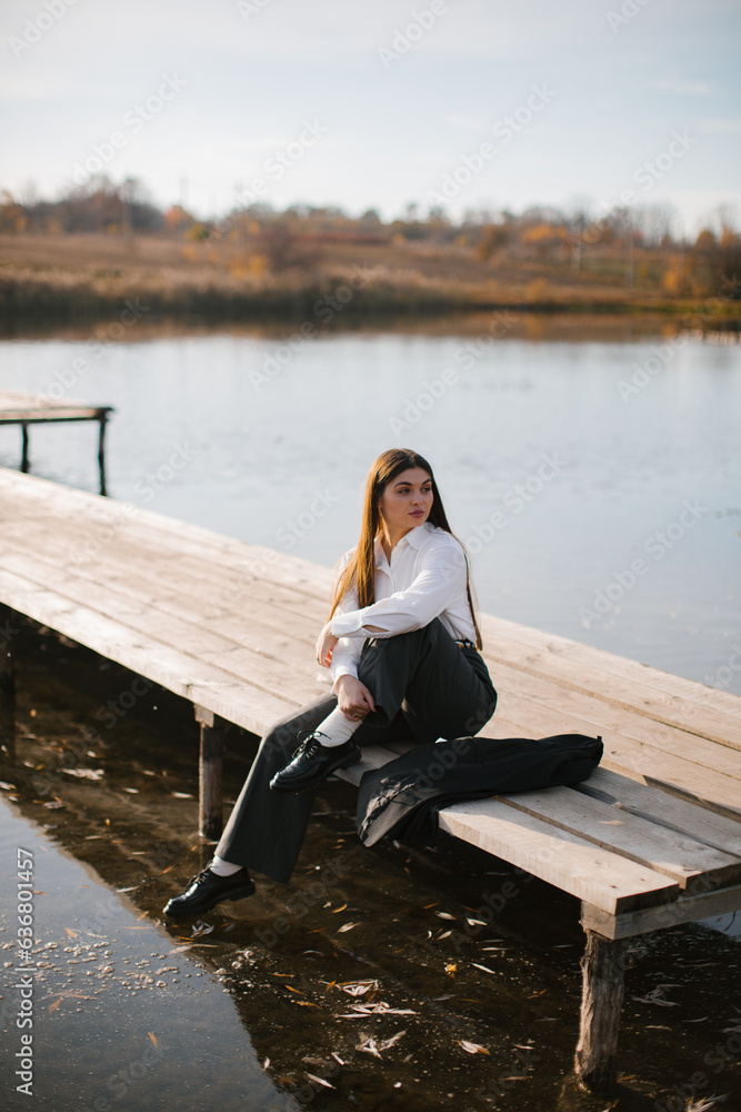 A woman with long hair in a white shirt and grey trousers is sitting on a wooden masonry near the lake in autumn. 