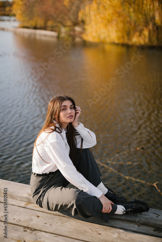 A woman with long hair in a white shirt and grey trousers is sitting on a wooden masonry near the lake in autumn. 